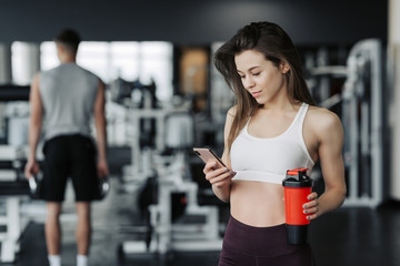 Beautiful young sportswoman in sportswear and earphones is holding a bottle of water, listening to music using a phone and smiling, in the gym.