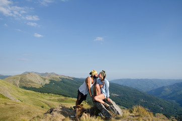 a couple is kissin on top of a mountains with their dog sit at their feet, and the landscape in background