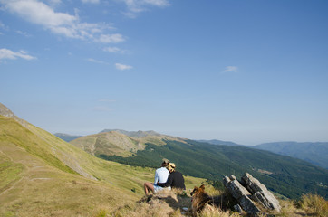 a couple is hugging on top of a mountains with their dog sit at their feet, and the landscape in background