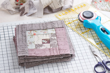 Patchwork log cabin blocks on craft mat, stack of blocks, sewing accessories on white wooden surface