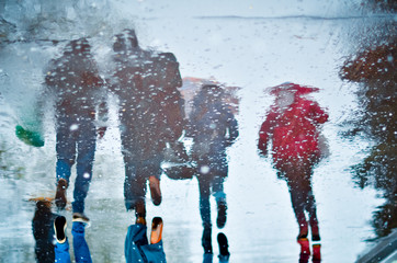 Blurry reflection in a puddle of four walking people on wet city street during rain and snow. Mood concept
