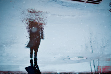 Blurry reflection in a puddle of alone walking person on wet city street during rain and snow. Mood concept - obrazy, fototapety, plakaty