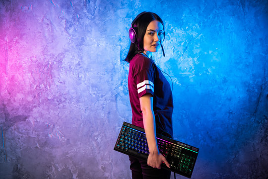 Beautiful Friendly Pro Gamer Streamer Girl Posing With a Keyboard in Her Hands, Wearing Glasses. Attractive Geek Girl with Cool Neon Retro Colors in Background.