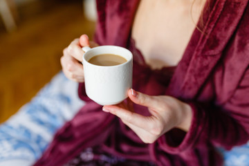 Morning coffee. Woman holding white Cup with black coffee sitting on bed in hotel. Young girl enjoys hot invigorating coffee in the morning