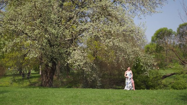 Young traveler pregnant woman walking, running, turning around and enjoys her leisure free time in a park with blossoming sakura cherry trees wearing a summer light long dress with flower pattern
