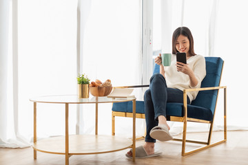 Young beautiful Asian woman relaxing in living room at home, reading books, drinking coffee and using phone in the morning with happy feeling