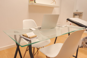 Interior of minimalist office with white walls, wooden floor, wooden and glass computer table and white bookcase with folders. Physiotherapy consultation room