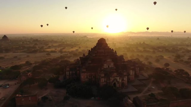 Ancient Buddhist temple in Bagan, Myanmar with dozens of hot air balloons in background during sunrise. Orbital temple shot.