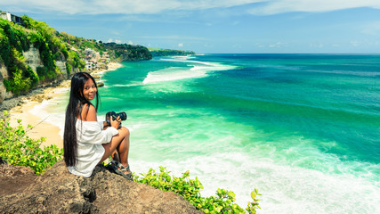Happy woman with camera in hand taking photo of beautiful nature landscape from top of mountain cliff in Bali. Active lifestyle, travel vacation concept