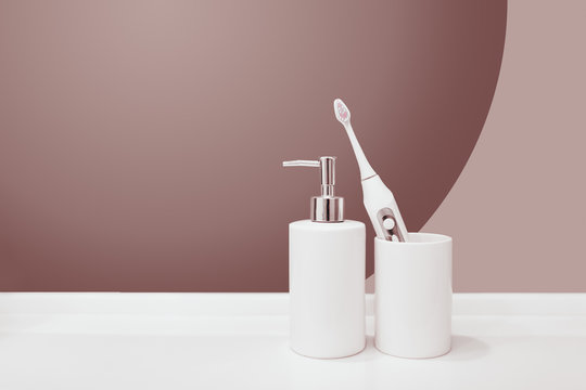 White ceramic bathroom accessories dispenser for soap and a glass with a tooth electric brush isolated on purple pastel background. copy space blank