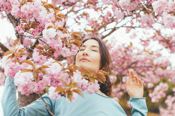 Fototapeta premium Portrait of beautiful brunette woman with blooming Japanese cherry tree (Prunus serrulata) - girl inhales the scent of flowers with closed eyes - spring, nature and beauty concept