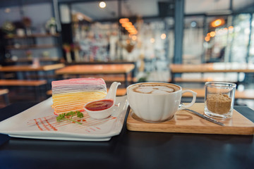 Rainbow crape cake and strawberry sauce on white plate with a cup of hot coffee on dark wooden table
