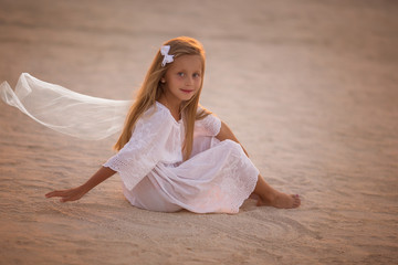 Fototapeta na wymiar Beautiful little girl in white dress sitting on the sand in the desert at sunset.