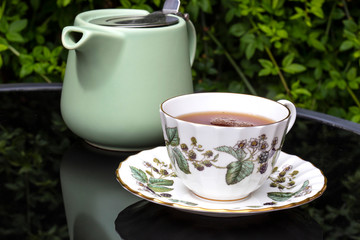 Cup of tea in a china teacup and saucer with a pastel green teapot reflected on a glass patio table outdoor