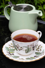 Cup of tea in a china teacup and saucer with a pastel green teapot reflected on a glass patio table outdoor