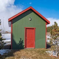 Square Small wooden storage shed with a lamp on the green wall above the red door