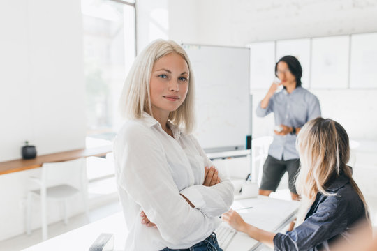 Confident blonde girl in blouse standing with arms crossed in office with big flipchart. Indoor portrait of serious female manager and her colleagues drinking coffee on background.