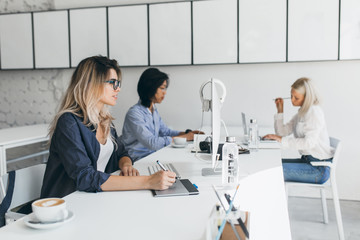 Indoor portrait of young web-developers of international company having coffee break. Blonde designer working with tablet while her asian colleague typing on keyboard.