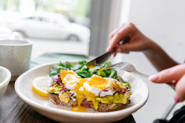 Young woman eat breakfast with knife and fork, Morning in cafe. Healthy breakfast with wholemeal bread toast with avocado, poached egg with green salad