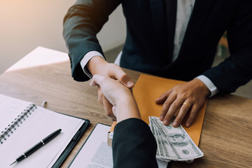 Two corporate businessmen shaking hands while one man places money on document in office room with corruption concept.