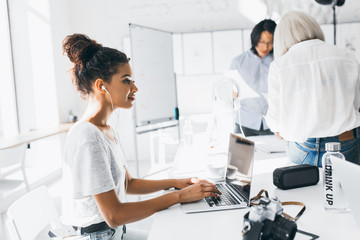 Portrait of relaxed freelance worker listening music in headphones and working with laptop. Portrait of young specialists of international corporation spending time in light cozy office.