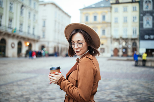 Young stylish woman in hat and coat drinking coffee to go in a city street