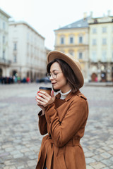 Young stylish woman in hat and coat drinking coffee to go in a city street