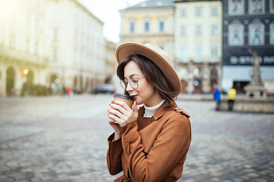 Young stylish woman in hat and coat drinking coffee to go in a city street