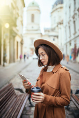 Young pretty woman use phone while walking to work drinking coffee on morning commute in city