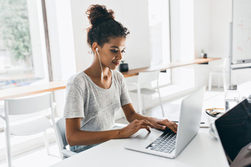 Romantic african girl with trendy hairstyle sitting at her workplace and analysing data. Indoor portrait of black female student working with laptop before exam.