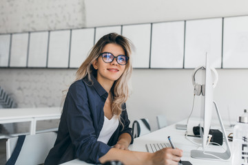 Pleased blonde young woman posing at her workplace and holding stylus. Indoor portrait of female...