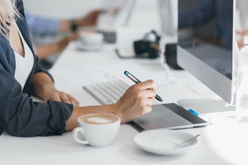 Portrait of freelance web-designer drinking coffee at workplace and holding stylus. Lightly-tanned...