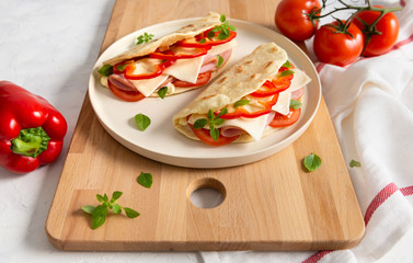 Italian piadina with cheese, tomato, ham, red pepper and basil on a white plate on a white wooden table. Side view, copy space