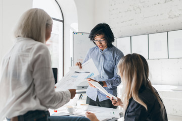 Young chinese man discussing new goals with cowokers and reading documents. Indoor portrait of busy students spending time in lecture hall while preparing for test.