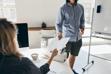 Young man in blue shirt and shorts carrying documents, walking by colleague's table. Indoor portrait of blonde girl drinking coffee in office and looking at flipchart.
