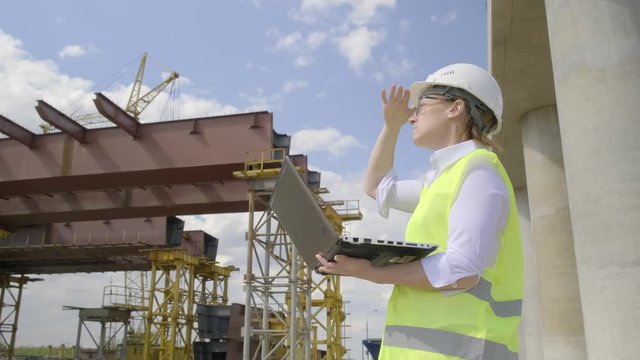 Portrait of engineer woman working on laptop on background of construction overpass on sunny day.