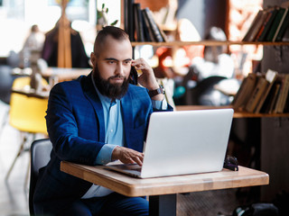 Fototapeta premium young bearded businessman at work in the office. A man sits at a laptop talking through a mobile phone. telephone conversations in coworking