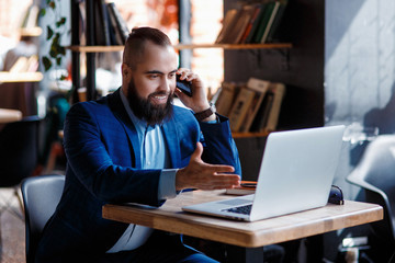 Fototapeta premium young bearded businessman at work in the office. A man sits at a laptop talking through a mobile phone. telephone conversations in coworking