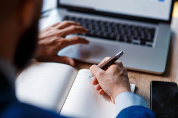 businessman in the office makes notes in a notebook. A student works in a laptop behind a laptop. close-up of computer, hands, and notebook