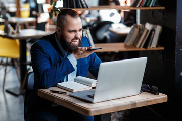 Fototapeta premium Serious bearded businessman conducts telephone conversations on a mobile phone at the computer. Unhappy man scolds on the phone. Emitations of rage in the office at the manager