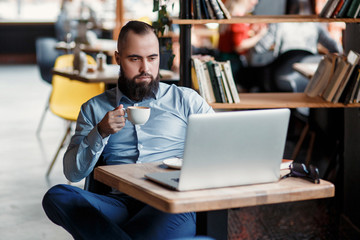 Fototapeta premium Young serious bearded businessman working on computer at table,drinking coffee.Man analyzes information, data, develops business plan. Freelancer, entrepreneur.Online marketing, education, e-learning