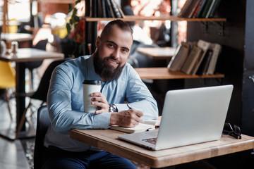 Fototapeta premium Young serious bearded businessman working on computer at table,drinking coffee.Man analyzes information, data, develops business plan. Freelancer, entrepreneur.Online marketing, education, e-learning