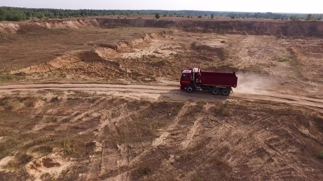 Aerial for the red big truck driving in ground quarry with flying dust behind it, industry concept. Scene. Empty lorry moving on country road with green forest on the background.