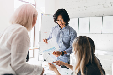 Fototapeta premium Asian male office worker with wristwatch holding documents with diagrams while talking with female colleagues. Indoor portrait of young executive in glasses discussing new goals.