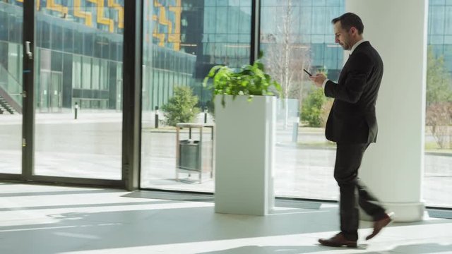 Full length tracking left shot of middle aged businessman in formal suit text messaging on cell phone while walking to work along entrance hall in modern business center