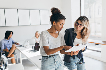 Slim black girl in jeans explaining something to european female colleauge while asian man talking with fair-haired young lady. Portrait of managers of international company solving work problems.
