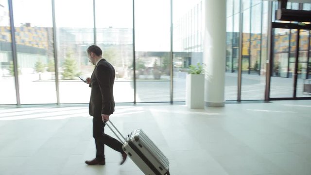 Tracking left shot of businessman in black formal suit texting on cell phone while walking with trolley suitcase across airport or hotel hall