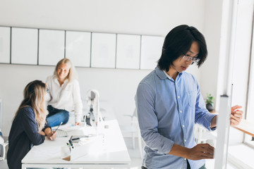 Fototapeta premium Blonde girl in denim pants sitting on table in office and talking with friend. Indoor portrait of young female secretary chilling at workplace and focused asian man on foreground.