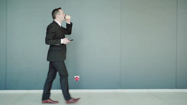 Tracking right side view of middle aged businessman in formal suit walking along corridor, text messaging on cell phone and drinking coffee from paper cup on the move