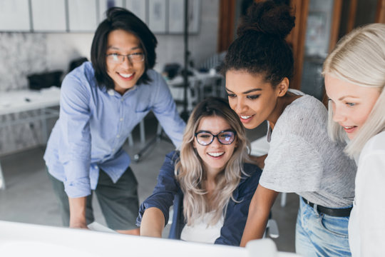 Close-up portrait of freelance it-specialists looking at laptop screen with smile. Asian programmer in glasses helping to mulatto girl in white t-shirt with her project.